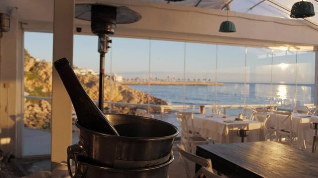 Empty seaside restaurant bathed in sunset light during the golden hour. A bottle of champagne or cava in an ice bucket near the table. Mediterranean interior, terrace, local cuisine, Sitges.