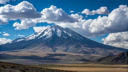 Fototapeta premium This stunning image captures a snow-capped mountain towering against a vibrant blue sky dotted with fluffy white clouds. The landscape features a wide,