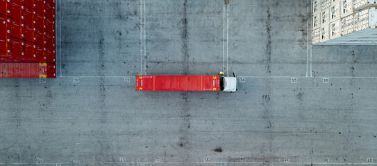 Aerial top-down view of a truck with a red shipping container parked in an industrial freight yard,...