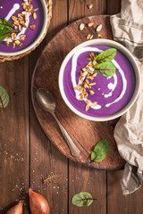 Two bowls of vibrant purple sweet popato soup garnished with a swirl of cream, toasted almonds, and fresh herbs, resting on a rustic wooden board.