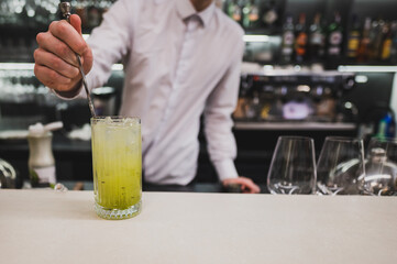 A bartender stirs a refreshing green cocktail in a tall glass at a modern bar. The atmosphere is lively, with a variety of bottles in the background and elegant glassware nearby.