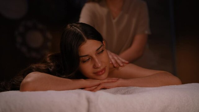 Close-up portrait of beautiful young woman enjoying moment of wellness and pampering, getting massage in beauty parlor with dark interior. Masseuse giving relaxing back massage to female client.
