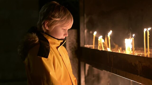 A beautiful boy looks at a group of thin burning candles in a church. A parishioner lights a candle after a church service in an Orthodox or Catholic church.Christian Christmas or Easter with children