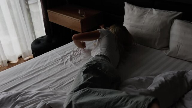 Blonde girl in white t-shirt and jeans with long wavy hair falls on her bedroom bed and cries