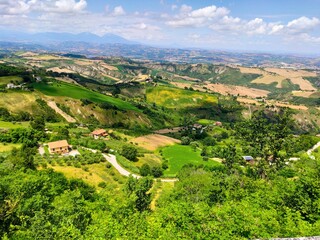 Obraz premium A vast panoramic view of the Italian countryside, featuring lush, rolling hills, cultivated fields, and scattered houses, all under a bright, partly cloudy sky.