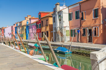 Italy, Burano. Canal and colorful buildings the island is known for. Mooring posts along the canal. Small boats moored to the sides.