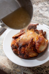 A view of a whole rotisserie chicken on a plate, featuring the pouring of gravy from a saucepan.
