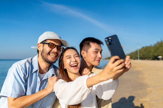 Happy man and woman using mobile phone during travel nature the sea on summer beach holiday vacation. Asian man enjoy and fun outdoor lifestyle using smartphone taking selfie with friends at the beach