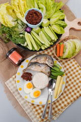 A meal setup featuring a cup of thai chili paste, a plate with two pieces of cooked fish, white rice, boiled egg and vegetables on  wooden tray