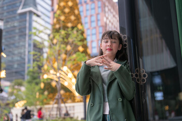 A young girl in a green coat posing gracefully in a festive urban setting, with a glowing Christmas tree and sparkling lights in the background, capturing a serene and joyful holiday moment.