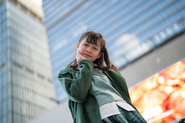 Fototapeta premium A confident young woman in a green coat standing outdoors, looking upward against the backdrop of a modern glass building, symbolizing ambition, contemporary style, and urban living.