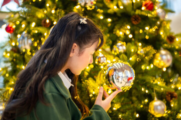 Naklejka premium A young girl in a green coat admiring a reflective ornament on a beautifully decorated Christmas tree, surrounded by glowing lights and festive holiday decorations, capturing a magical moment.