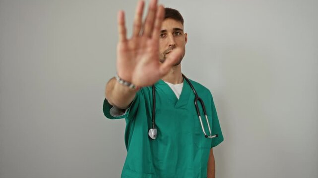 Young doctor in green scrubs with stethoscope gesturing stop with hand against white background, expressing caution and seriousness, standing with focus on medical profession and care