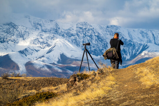 Person photographing scenic snowy mountains using camera on tripod under cloudy sky