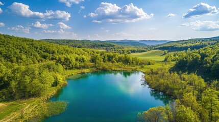 Fototapeta premium Vista aérea de un lago rodeado de verdes colinas y un bosque bajo un cielo azul con nubes esponjosas
