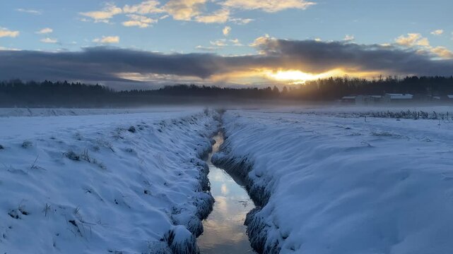 Snow-covered field with a stream and mist during sunrise.