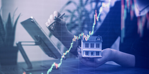 Professionals Reviewing Data with Miniature House Model and Stock Market Candlestick Chart Overlay in a Business Setting