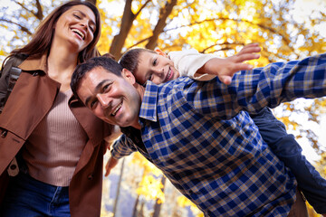 Fototapeta premium Smiling father gives his young son a piggyback ride on his back in autumn park. Mother is smiling. Family in the park.