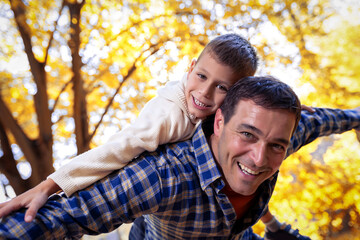 Smiling father gives his young son a piggyback ride on his back in autumn park.