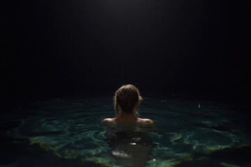 a woman in a dark environment, partially submerged in water with their back to the camera. The background is almost entirely black, creating a loneliness and fear.