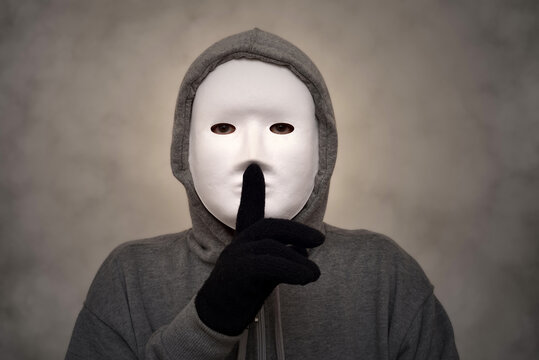 Unrecognizable person wearing hoodie, white mask and black gloves holding a finger to their lips and showing a symbol of silence. Smoke or steam effect as a background.