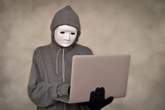 Computer hacker wearing hoodie, white mask and black gloves using a laptop computer for cyber attack. Smoke or steam effect as a background. Hacker attack and cyber security concept. Selective focus. 