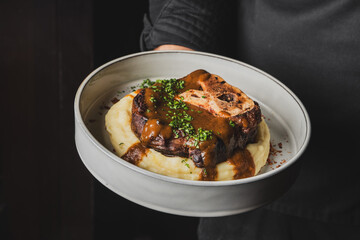 A chef holds a plate featuring a succulent piece of meat with rich gravy, served on a bed of creamy mashed potatoes, garnished with fresh herbs. Perfect for gourmet food showcases.