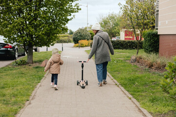 Mother supports her daughter while she learns to ride a scooter outdoors, close-up. The concept of...