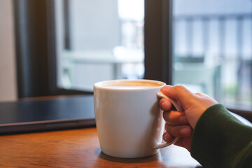 A hand holding a cup of coffee with laptop on the table