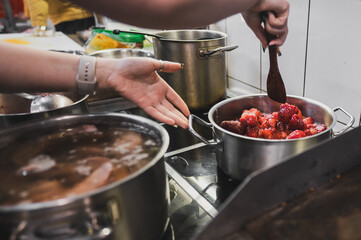 A close-up of a kitchen scene with hands stirring a pot of red fruit. Other pots on the stovetop indicate cooking activity, creating a vibrant and lively culinary atmosphere.