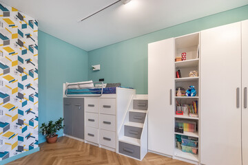 A children's room with a modern interior. High bed with stairs and built-in storage drawers. Spacious wardrobe with shelves. Multicolored print on the wall.
