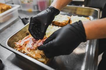 A close-up of a chef's hands in black gloves assembling a sandwich. Fresh ingredients like bacon and cabbage are being layered on toasted bread in a commercial kitchen setting.