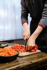 Unrecognizable man in a sweater and apron skillfully cuts red bell peppers on a wooden cutting board
