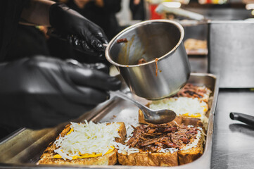 A chef in black gloves prepares a delicious sandwich, layering shredded cheese and pulled meat onto slices of toasted bread in a bustling kitchen setting.