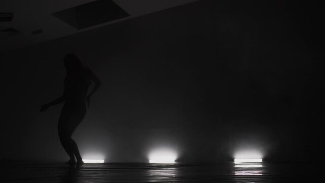 Modern ballet dancer performing on dark stage. Silhouette of female performer with smoke in the air.