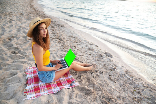 Work from anywhere in the world. Side view of young female freelancer in straw hat working on laptop while sitting on tropical sandy beach at sunset. Isolated laptop screen green screen