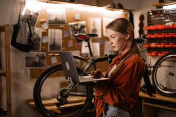 Fototapeta premium Cute blonde woman using a laptop computer in bicycle repair shop. Running a business and start-up.