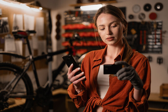 Woman mechanic repairing bicycle while standing with credit card and mobile phone.