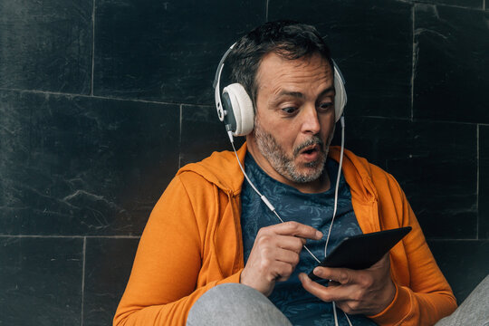 middle aged man with headphones and surprised gesture sitting on the street using digital tablet