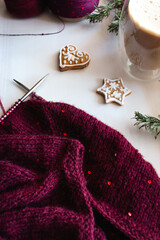 Red knitted sweater with sequins and knitting needles on white background with gingerbread cookies, coffee cup and pine tree branches. Christmas decoration. Sequin pattern in needlework with coffee