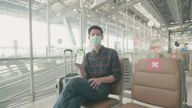 A traveler sits at the airport, wearing a face mask and holding a smartphone. The contemporary airport design and safety decals reflect the importance of health precautions during travel.
