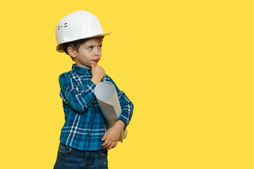 Thinking little kid boy engineer or architect in a protective helmet holding construction plan over yellow background.