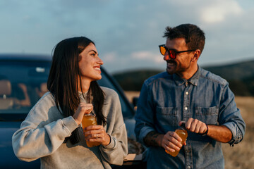 Relaxed couple sharing a pizza and juices by their car with a stunning meadow sunset in the...