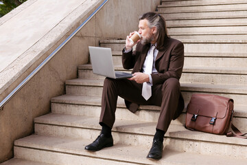 elegant middle age male entrepreneur with beard in formal suit sitting on urban stairway drinking takeaway coffee and working online on laptop in city, concept of business and middle age man lifestyle