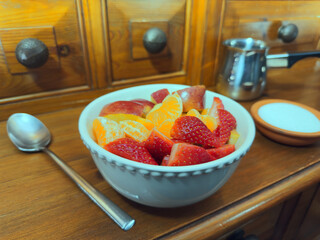 A bowl of chopped fruit - orange, apple and strawberries - on a pine kitchen dresser, next to sugar, cream and a spoon.