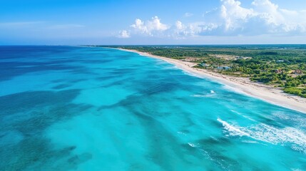Obraz premium Aerial perspective of vibrant turquoise ocean waves meeting a serene sandy shore under a clear blue sky with scattered clouds.