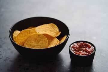 Round nachos in black ceramic bowl on concrete background with red salsa