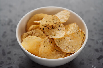 White bowl with organic potato chips sprinkled with fresh ground black pepper on concrete background