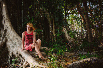 Portrait of a girl in a tropical jungle. Standing at the huge roots of a tree in a red dress