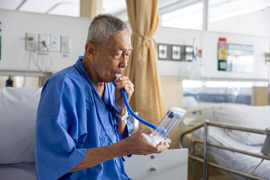 Senior asian man using incentive spirometer device for breathing exercise rehabilitation to recover from lung cancer after surgery due to heavy smoking in hospital bed